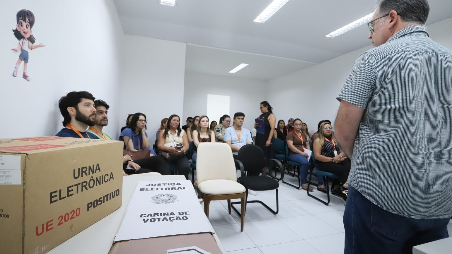 A foto mostra uma sala de aula com pessoas sentadas em carteiras. Em primeiro plano há uma caixa sinalizada como 